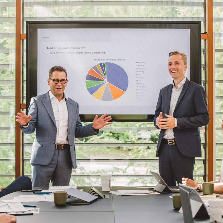 Two business professionals in suits stand in front of a presentation screen displaying a colorful pie chart. One gestures while speaking, and the other smiles, as colleagues sit at a conference table with laptops and documents.